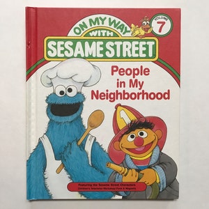 May include: A red, white and blue Sesame Street book titled "People in My Neighbourhood" featuring Cookie Monster and Elmo. The book is part of the "On My Way with Sesame Street" series and is volume 7.