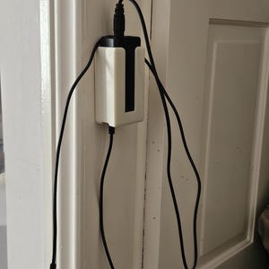 Könnte beinhalten: Eine weiße Kunststoff-Wandhalterung, die ein schwarzes, rechteckiges Netzteil sichert. Das Netzteil hat ein schwarzes Stromkabel, das oben herausführt, und ein langes schwarzes Ladekabel, das unten in losen Schlaufen herabhängt.