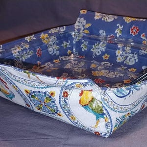 May include: A fabric casserole dish cover with a clear glass dish inside. The cover features a white paisley pattern with blue, yellow, and red accents. The interior is a blue floral print.