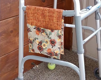 May include: A silver metal walker with gray rubber hand grips and two yellow tennis balls attached to the front legs. A fabric pouch with an orange textured top and a floral sunflower print bottom hangs from the side frame of the walker.