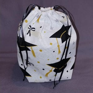 May include: A white fabric drawstring bag featuring a repeating pattern of black graduation caps with gold tassels, black bows, and small gold and silver stars and dots. The bag is cinched at the top with thin black ribbon drawstrings on both sides.