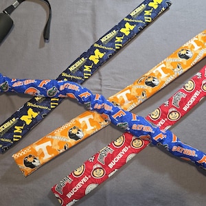 May include: A collection of four fabric-covered items, each with a different college team theme. The items are in blue, orange, and red, with team names and logos printed on them. A black handle is attached to one of the items.