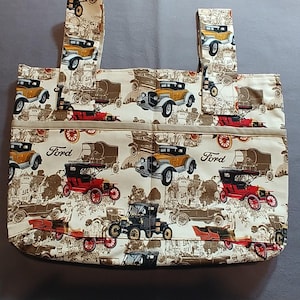 May include: A tote bag featuring a cream-colored fabric with a repeating pattern of vintage Ford automobiles in yellow, red, and black. The design includes the Ford logo printed in black script, two fabric handles, and a horizontal front pocket.