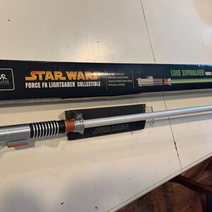 May include: A Star Wars Force FX Lightsaber collectible, featuring a silver and black hilt with orange accents and a glowing white blade. The lightsaber is displayed on a black stand with the Star Wars logo, next to its black box.