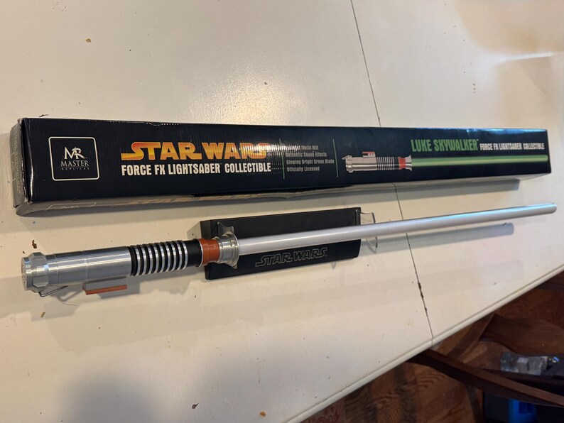 May include: A Star Wars Force FX Lightsaber collectible, featuring a silver and black hilt with orange accents and a glowing white blade. The lightsaber is displayed on a black stand with the Star Wars logo, next to its black box.