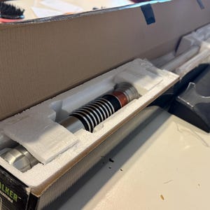 May include: A Luke Skywalker collectible lightsaber in its original packaging. The lightsaber features a silver handle with black and white details, and is displayed in a cardboard box with foam inserts.