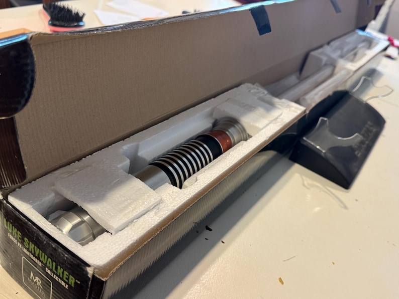 May include: A Luke Skywalker collectible lightsaber in its original packaging. The lightsaber features a silver handle with black and white details, and is displayed in a cardboard box with foam inserts.