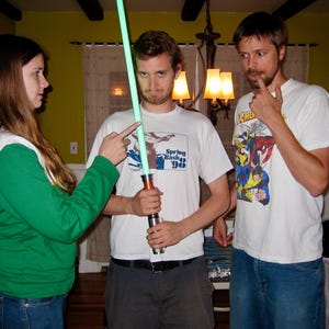 May include: Three people are gathered, with one holding a green lightsaber. The person in the center wears a white t-shirt with the text "Spring Bash '98". The other two people are pointing at the lightsaber.