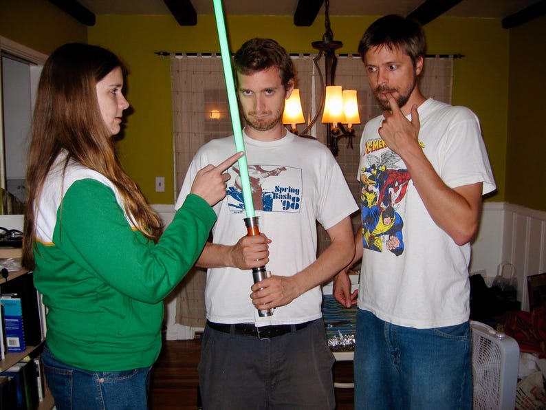 May include: Three people are gathered, with one holding a green lightsaber. The person in the center wears a white t-shirt with the text "Spring Bash '98". The other two people are pointing at the lightsaber.
