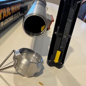 May include: A disassembled Star Wars FX Lightsaber Collector's Edition. The silver hilt is next to a black battery compartment and a silver cap. The box with the Star Wars logo is partially visible.