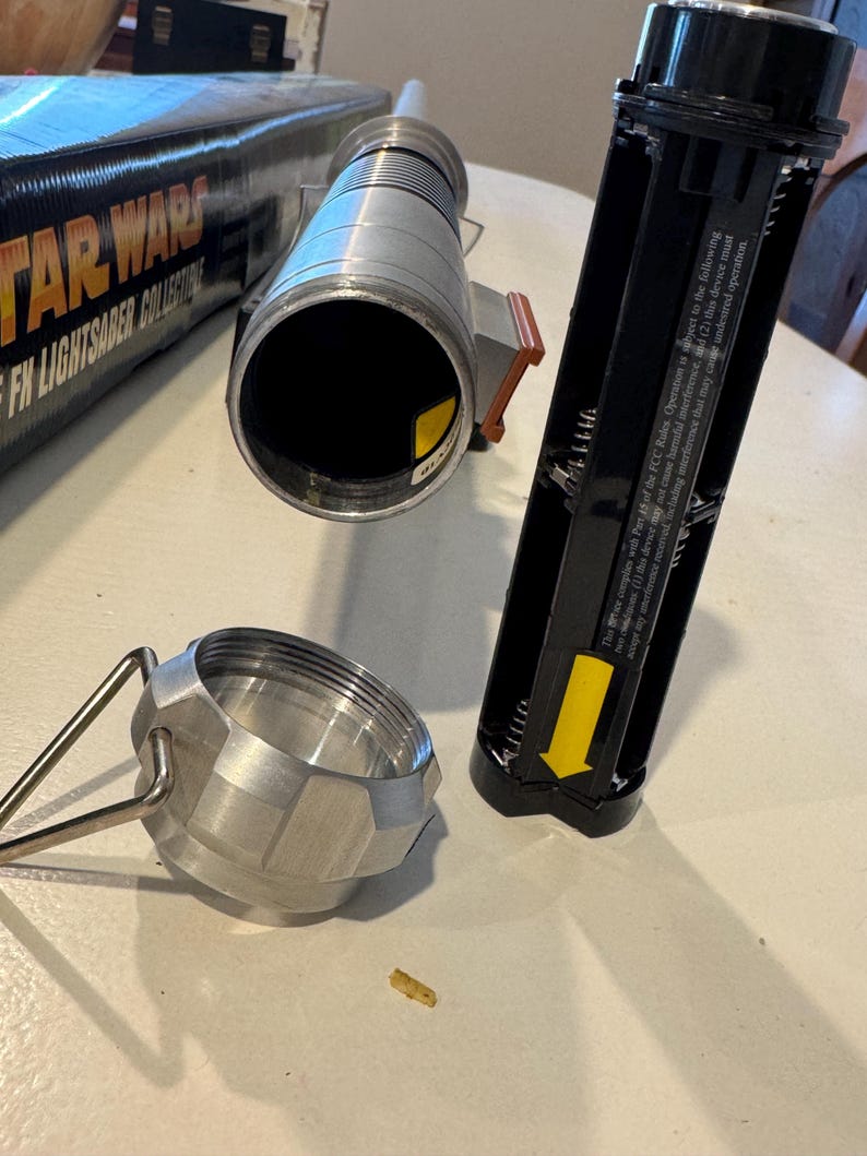 May include: A disassembled Star Wars FX Lightsaber Collector's Edition. The silver hilt is next to a black battery compartment and a silver cap. The box with the Star Wars logo is partially visible.