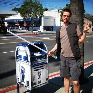 May include: A blue and white United States Postal Service mailbox, designed to resemble the Star Wars character R2-D2, with a lightsaber extending from it. A person stands next to the mailbox, holding the lightsaber.