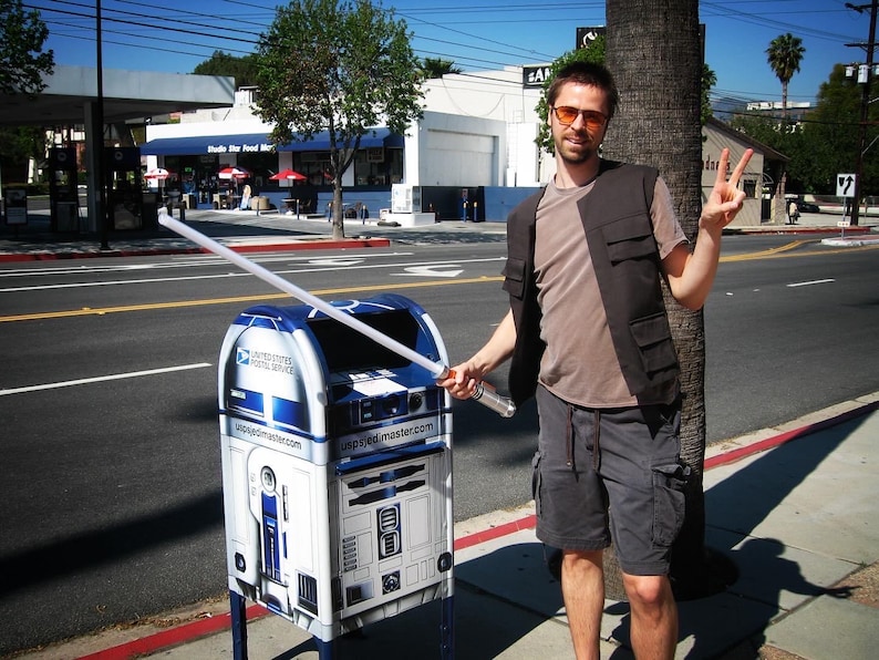 May include: A blue and white United States Postal Service mailbox, designed to resemble the Star Wars character R2-D2, with a lightsaber extending from it. A person stands next to the mailbox, holding the lightsaber.