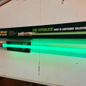 May include: A Star Wars Luke Skywalker Force FX Lightsaber collectible. The lightsaber has a green blade and a silver and black hilt. The lightsaber is displayed with its black box, which has the Star Wars logo and the words "Luke Skywalker Force FX Lightsaber Collectible."