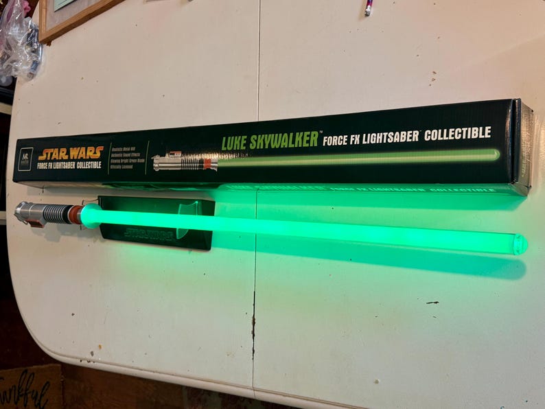 May include: A Star Wars Luke Skywalker Force FX Lightsaber collectible. The lightsaber has a green blade and a silver and black hilt. The lightsaber is displayed with its black box, which has the Star Wars logo and the words "Luke Skywalker Force FX Lightsaber Collectible."