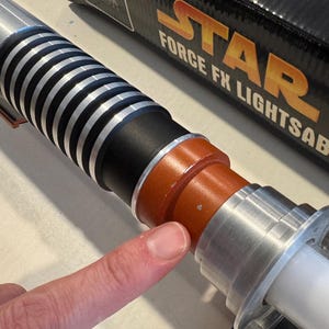 May include: A silver and black lightsaber with a white blade. The lightsaber has a ribbed handle and orange and black accents. The box in the background reads "STAR WARS FORCE FX LIGHTSABER".