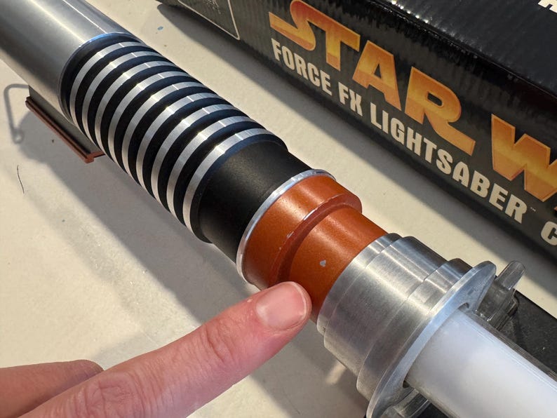 May include: A silver and black lightsaber with a white blade. The lightsaber has a ribbed handle and orange and black accents. The box in the background reads "STAR WARS FORCE FX LIGHTSABER".