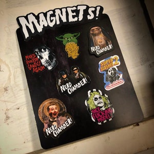Puede incluir: Una pizarra magnética negra con un título blanco "MAGNETS!" y varios imanes coloridos con personajes de películas y programas de televisión populares. Los imanes incluyen una criatura de la laguna negra, un gremlin, un personaje de la película "Beetlejuice" y un personaje de la película "Super Slasher 2".