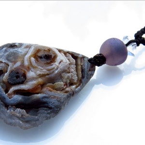 K&ouml;nnte beinhalten: Eine einzigartige Anh&auml;nger Halskette mit einem Naturstein mit geodenartigem Innenraum. Der Stein ist haupts&auml;chlich grau, braun und wei&szlig;, mit einer schwarzen Kordel und einer lavendelfarbenen Perle. Handgefertigter Schmuck.