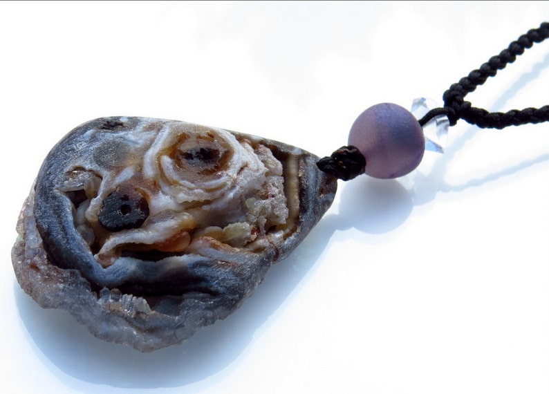 K&ouml;nnte beinhalten: Eine einzigartige Anh&auml;nger Halskette mit einem Naturstein mit geodenartigem Innenraum. Der Stein ist haupts&auml;chlich grau, braun und wei&szlig;, mit einer schwarzen Kordel und einer lavendelfarbenen Perle. Handgefertigter Schmuck.