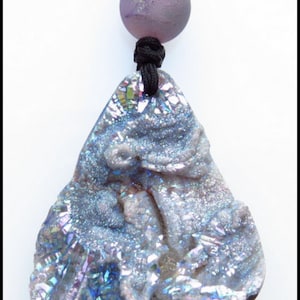 K&ouml;nnte beinhalten: Halskette mit einem tropfenf&ouml;rmigen, schillernden Steinanh&auml;nger. Der Anh&auml;nger h&auml;ngt an einer schwarzen Kordel, mit einer lila Perle und einem klaren Kristallakzent. Der Stein hat Schattierungen von Blau, Lila und Silber. Ein einzigartiges Schmuckst&uuml;ck.