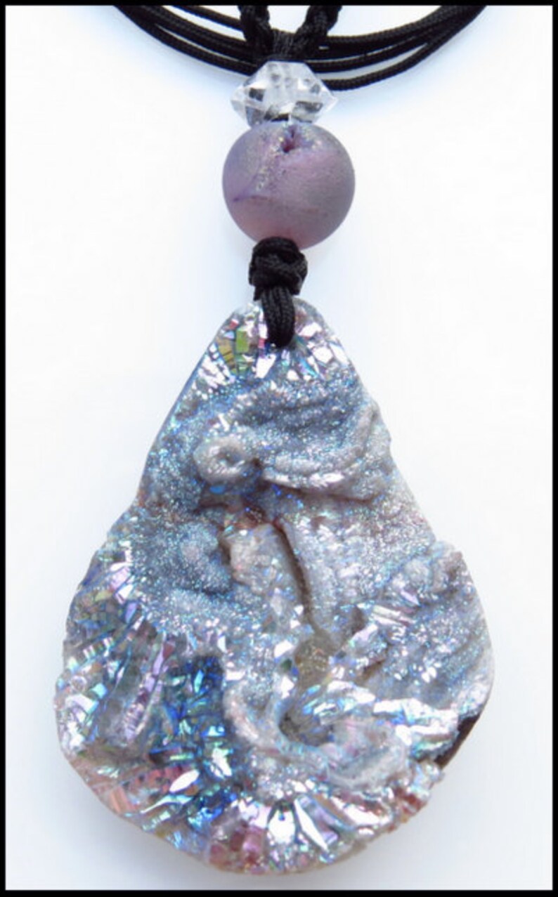 K&ouml;nnte beinhalten: Halskette mit einem tropfenf&ouml;rmigen, schillernden Steinanh&auml;nger. Der Anh&auml;nger h&auml;ngt an einer schwarzen Kordel, mit einer lila Perle und einem klaren Kristallakzent. Der Stein hat Schattierungen von Blau, Lila und Silber. Ein einzigartiges Schmuckst&uuml;ck.