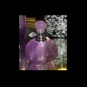 Puede incluir: Un frasco de perfume de amatista púrpura facetada con cuello y tapón plateados. El frasco tiene forma redondeada y se exhibe sobre una superficie reflectante. Un objeto estampado se encuentra en el fondo.