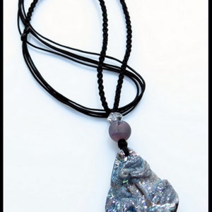 K&ouml;nnte beinhalten: Eine schwarze Kordelkette mit einem Anh&auml;nger. Der Anh&auml;nger ist ein gro&szlig;er, unregelm&auml;&szlig;ig geformter Stein mit einem geschnitzten Design und schillernden Farben. Eine lila Perle und eine klare Kristallperle befinden sich ebenfalls an der Halskette.