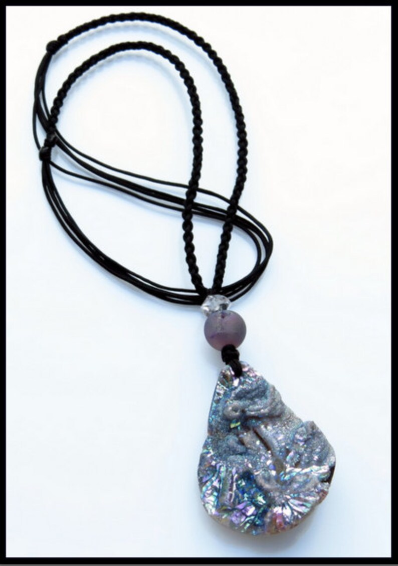 K&ouml;nnte beinhalten: Eine schwarze Kordelkette mit einem Anh&auml;nger. Der Anh&auml;nger ist ein gro&szlig;er, unregelm&auml;&szlig;ig geformter Stein mit einem geschnitzten Design und schillernden Farben. Eine lila Perle und eine klare Kristallperle befinden sich ebenfalls an der Halskette.