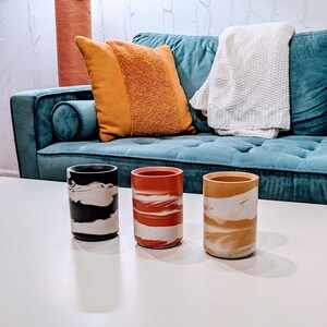 May include: Three decorative concrete candle holders with a marbled design. The candle holders are black, red, and yellow.