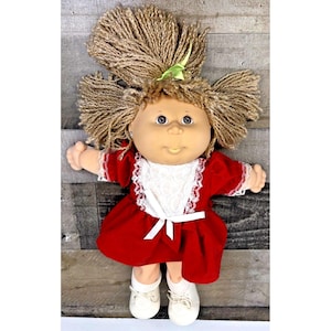 Pode incluir: Uma boneca com cabelo de lã castanho-claro em marias-chiquinhas com uma fita verde. A boneca tem rosto redondo, olhos azuis e nariz pequeno. Veste um vestido vermelho com um babador de renda branca, um laço branco na cintura e sapatos.