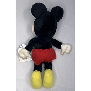 Puede incluir: Un juguete de peluche Mickey Mouse negro con pantalones cortos rojos y zapatos amarillos. El juguete est&aacute; de pie sobre una superficie blanca y tiene una etiqueta adherida a su brazo.