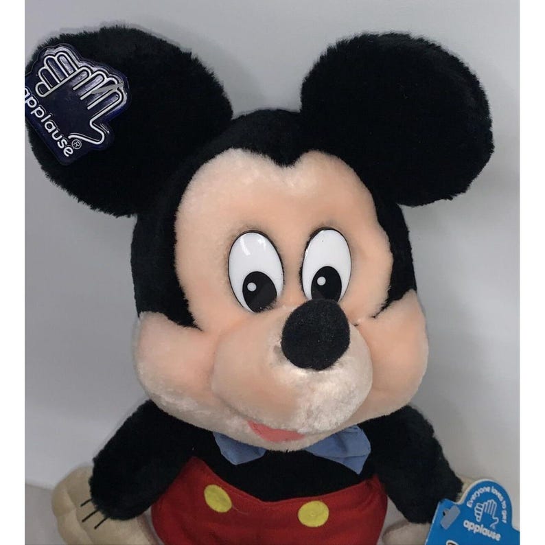 Puede incluir: Un juguete de peluche de Mickey Mouse con orejas negras, nariz negra y camisa de lunares rojos y amarillos. El juguete tiene una pajarita azul y el texto "applause" en la etiqueta.