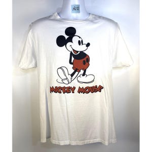 May include: White t-shirt featuring a classic Mickey Mouse graphic. The design includes a smiling Mickey in his signature red shorts, black ears, and white gloves and shoes. The words "Mickey Mouse" are printed in red below the character.