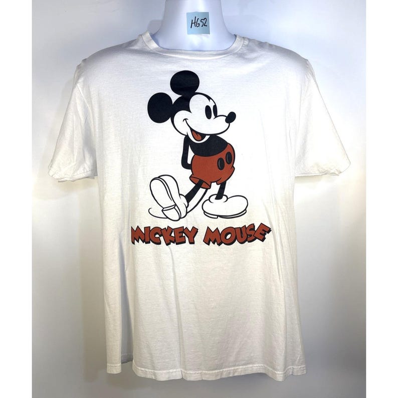 May include: White t-shirt featuring a classic Mickey Mouse graphic. The design includes a smiling Mickey in his signature red shorts, black ears, and white gloves and shoes. The words "Mickey Mouse" are printed in red below the character.
