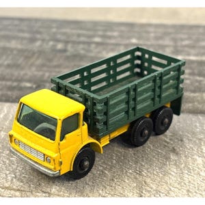Puede incluir: Un camión de juguete vintage amarillo y verde. La cabina es amarilla con la palabra "Dodge", y la caja es verde. El camión tiene ruedas negras y un parachoques plateado. Un juguete clásico con un diseño detallado.