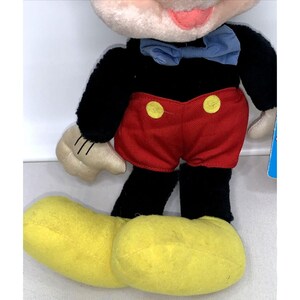 May include: A plush Mickey Mouse toy with black fur, red shorts with yellow buttons, and yellow shoes. The toy has a blue bow tie and a pink face.