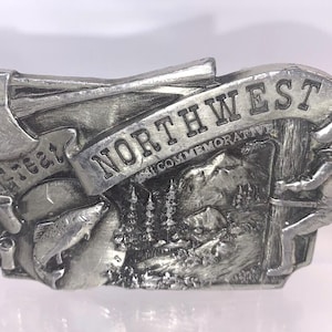May include: A silver belt buckle with a detailed design featuring a mountain scene, trees, and a figure holding a log. The buckle has the words "Great Northwest Commemorative" inscribed on it.