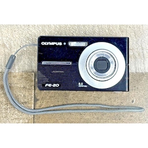 May include: Black Olympus FE-20 digital camera with a silver lens and a gray wrist strap. The camera has the Olympus logo and the text "8.0 Megapixels". The camera is rectangular and has a silver lens ring.
