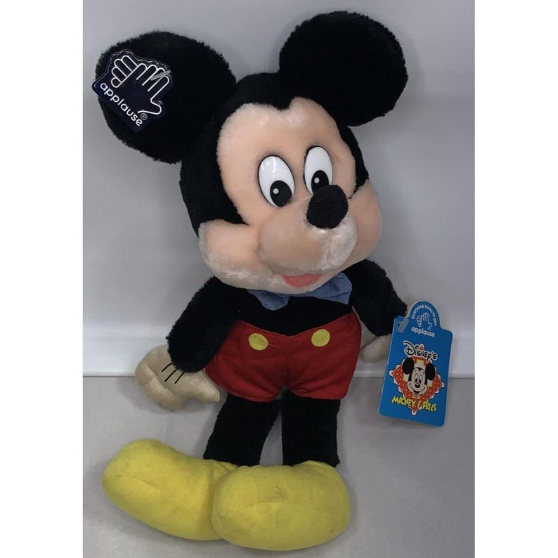 Puede incluir: Un juguete de peluche de Mickey Mouse que lleva pantalones cortos rojos con botones amarillos, zapatos negros y una pajarita azul. El juguete sostiene un objeto dorado en su mano derecha. El juguete tiene una etiqueta que dice "Disney's Mickey Mouse Clubhouse".