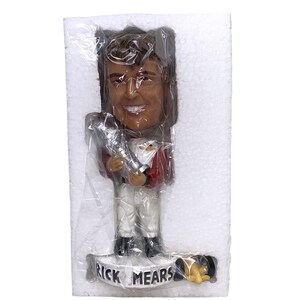 May include: A bobblehead figurine of a smiling man in a red and white racing suit, holding a trophy. The base of the figurine reads "RICK MEARS". The figurine is packaged in clear plastic and white foam.