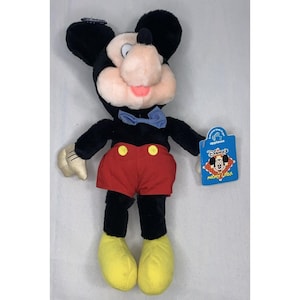 Puede incluir: Un juguete de peluche Mickey Mouse negro y rojo con una pajarita azul. El juguete lleva zapatos amarillos y tiene una etiqueta que dice "Disney's Mickey Mouse Clubhouse".