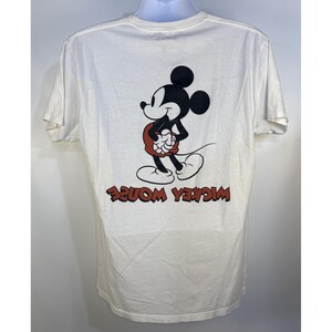 May include: White t-shirt featuring a graphic of Mickey Mouse in black, red, and white. The words "Mickey Mouse" are printed in red, in reverse, at the bottom of the shirt. The shirt has short sleeves and a crew neck.