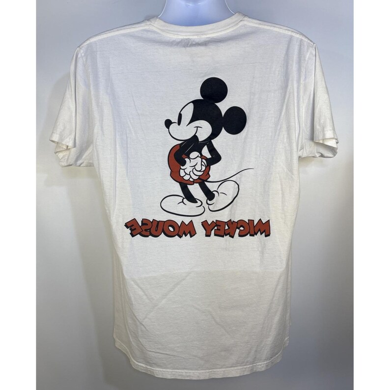 May include: White t-shirt featuring a graphic of Mickey Mouse in black, red, and white. The words "Mickey Mouse" are printed in red, in reverse, at the bottom of the shirt. The shirt has short sleeves and a crew neck.