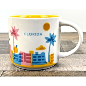May include: White ceramic mug with a yellow interior and a white handle. The mug features a colorful Florida skyline design with palm trees, buildings, and the word "FLORIDA". The mug is a standard size for coffee or tea.