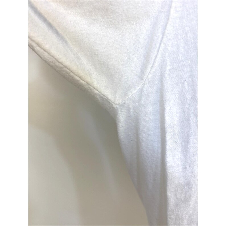May include: Close-up of a white V-neck shirt. The fabric appears to be a soft knit, with visible texture and slight discoloration under the arm. The neckline is a sharp V-shape.