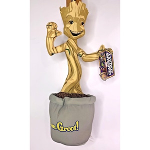 Puede incluir: Un juguete de peluche de Groot de Guardianes de la Galaxia. Groot está de pie en una maceta con las palabras "¡Yo soy Groot!" en la parte delantera. El juguete es marrón y verde y tiene una expresión feliz en su rostro.