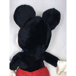 May include: Black plush Mickey Mouse toy with red shorts and white gloves. The toy is facing away from the camera, showing the back of its head.