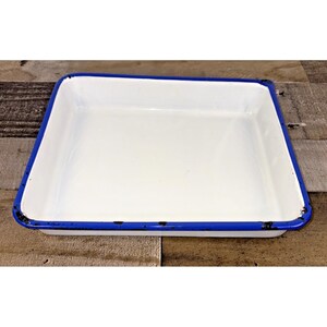 May include: A rectangular white enamel tray with a bright blue rim. The tray has a slightly distressed appearance, with some wear visible on the blue edge. The tray is empty and sits on a wooden surface.
