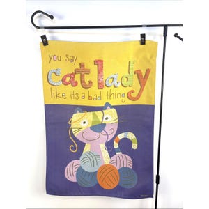Puede incluir: Una bandera de jardín morada y amarilla con un gato de dibujos animados sosteniendo ovillos de lana. El texto en la bandera dice "You say cat lady like it's a bad thing."